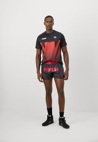 Schwarzes sportliches T-Shirt mit kurzen Ärmeln und rotem Grafikdruck, kombiniert mit schwarzen Shorts, die roten "VENUM"-Text und helle Akzente aufweisen. Schwarze Sneakers.
