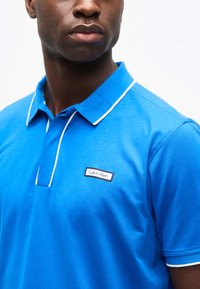 Homme portant un polo Calvin Klein bleu vif avec des bordures blanches sur le col et les manches, visible des épaules au menton.