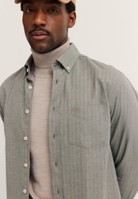 Mann trägt einen beigen Rollkragenpullover unter einem grau-grünen Hemd mit Knöpfen und einer beige Kappe vor einem hellen, einfarbigen Hintergrund.