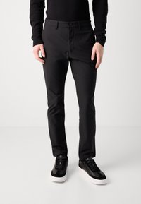 Pantalones negros hechos de una tela suave, con un corte ajustado y un diseño de frente plano. Combinados con zapatillas negras con suelas blancas.