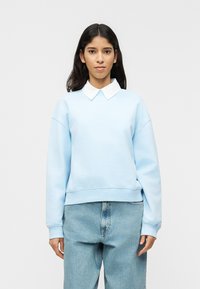 Γυναίκα με μακριά σκουρόχρωμα μαλλιά, ντυμένη με ανοιχτό γαλάζιο φούτερ πάνω από λευκό πουκάμισο με γιακά και φαρδιά μπλε τζιν, στέκεται μπροστά σε απλό φόντο.