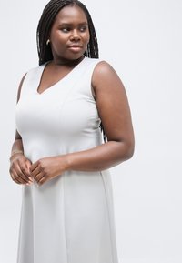 Γυναίκα με μακριά πλεγμένα μαλλιά που φοράει αμάνικο ανοιχτό γκρι φόρεμα, κοιτάζοντας προς τα πλάγια με τα χέρια της απαλά σταυρωμένα μπροστά.