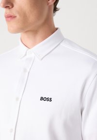 Fehér pólóing, textúrált anyagból készült. Gombos gallérral és fekete hímzett "BOSS" logóval rendelkezik a bal mellen.