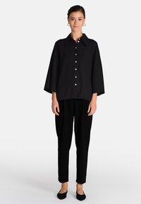 Camicia nera con colletto grande e maniche svasate, abbinata a pantaloni neri a sigaretta e scarpe basse, creando un look monocromatico.