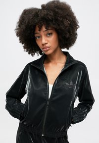 Juodos zip-up treniruotės striukės, pagamintos iš blizgaus audinio, su stovinčiu apykakle, įmegztomis rankogaliais ir subtiliu logotipo detaliu krūtinėje.