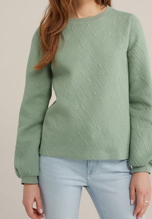 Frau trägt ein salbeigrünes, strukturiertes Langarmshirt mit Ballonärmeln und hellblaue Jeans vor einfachem Hintergrund.