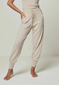 Beige gestrickte Jogginghosen mit einem elastischen Bund und Kordelzug, seitlichen Taschen und schmal zulaufenden Bündchen an den Knöcheln. Weicher Stoff.