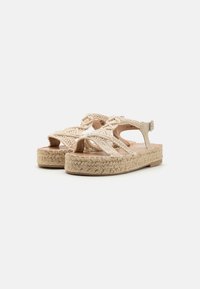 Sandalias de espadrille tejidas en color beige con tiras entrelazadas, suela plana de yute y cierre de hebilla. Presentan una textura natural y un diseño ligero.