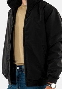 Zwarte zip-up jas met een staande kraag, elastische manchetten, voorzien van een subtiele logo op de borst en een gladde stof. Gecombineerd met een beige shirt.