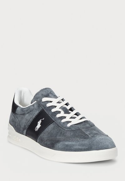 Grå mockasinsneaker med vita snören, med en mörkblå läderdetalj och logotyp på sidan. Rund tå och texturerad ovandel.