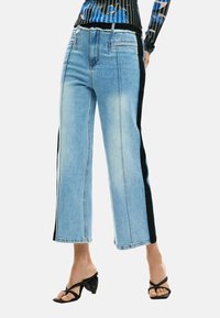 Hele sinised laia lõikega teksapüksid mustade külgpaneelidega, lõdva istuvusega ja narmendava äärega. Kanditud mustade kontsakingadega sandaalidega.