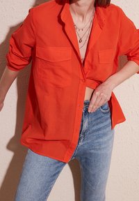 Femme portant une chemise orange vif déboutonnée avec des manches retroussées sur un jean bleu clair, ajustant sa taille avec des colliers superposés visibles.