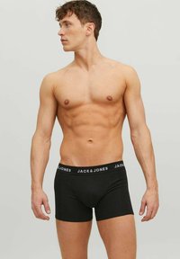 Shirtless man with toned muscles wearing black Jack & Jones boxer briefs, standing against a plain light gray background, looking left.
