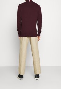 Bordeauxfarbener Strickpullover mit hohem Kragen und rot-weiß-blauem Streifen auf dem Rücken, kombiniert mit lockeren beigen Hosen und schwarzen Sneakern.