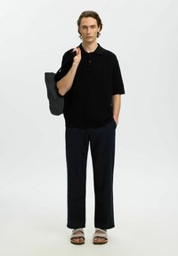 Jeune homme debout portant un polo noir, un pantalon foncé, des sandales, et une sacoche grise en bandoulière sur un fond uni.