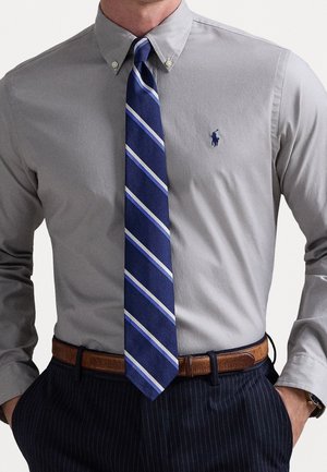 Homme portant une chemise grise à boutons, une cravate bleue rayée, une ceinture marron et un pantalon sombre à fines rayures, les mains dans les poches.