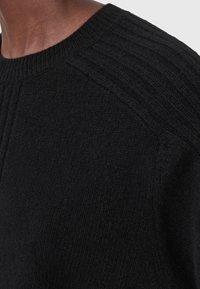 Primo piano di una persona che indossa un maglione nero lavorato a maglia con dettagli a costine sulle spalle e sul colletto.