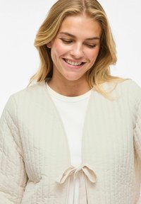 Giacca trapuntata di un beige chiaro con chiusura a legaccio, caratterizzata da una texture liscia e dettagli di cucitura sottili. Indossata sopra un top bianco.