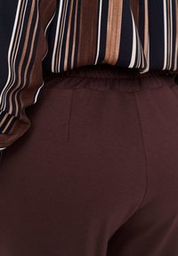 Persoon draagt donkerbruine broek met elastische tailleband en een ingestopt overhemd met verticale bruine, zwarte en witte strepen.