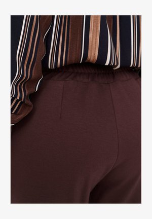 Persoon draagt donkerbruine broek met elastische tailleband en een ingestopt overhemd met verticale bruine, zwarte en witte strepen.