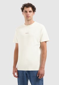 Crème katoenen T-shirt met korte mouwen, ronde hals en kleine "PURE PATH" print op de borst. Gecombineerd met blauwe denim jeans.