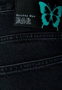 Černý denim s viditelnými švy, s etiketou obsahující "Bershka Man" a grafikou modré motýlice.