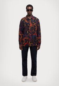 Camicia button-up con un motivo floreale scuro nei toni del rosso, viola e oro, abbinata a jeans dritti scuri e sneakers bianche.
