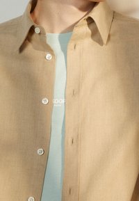 Lichtbruine blouse met knopen en een lichte textuur, voorzien van een kraag, witte knopen en een subtiele glans, gedragen over een lichtblauw shirt met ronde hals.