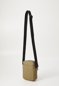 Geantă crossbody khaki, confecționată dintr-un material moale, cu o formă rectangulară, închidere cu fermoar și o curea texturată neagră cu branding.