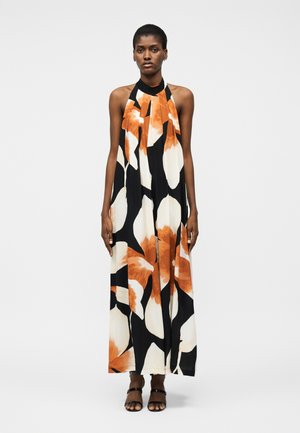 Γυναίκα όρθια, ντυμένη με ένα αμάνικο, μακρύ φόρεμα με λαιμόκοψη χιαστί, με μεγάλα μαύρα, λευκά και πορτοκαλί ανθικά σχέδια και μαύρα παπούτσια με ανοιχτά δάχτυλα.