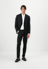 Traje negro con chaqueta ajustada y pantalones slim-fit, combinado con una camisa blanca y zapatos negros. Tejido suave y diseño moderno con detalles sutiles.