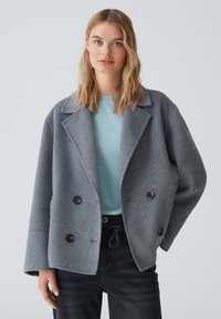 Doppeltknöpfige graue Wolljacke mit Kragen, großen Knöpfen und weiten Ärmeln, getragen über einem hellblauen Top und dunklen Jeans.