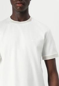Camiseta blanca de manga corta hecha de algodón, con acentos ribeteados en beige en el cuello y las mangas, que presenta un discreto logo "BOSS" en el pecho.