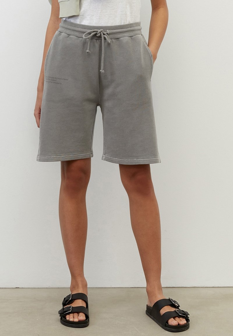 Shorts gris en mélange de coton avec une ceinture élastique, un cordon de serrage, des poches latérales et des détails de couture visibles. Portés avec des sandales noires.