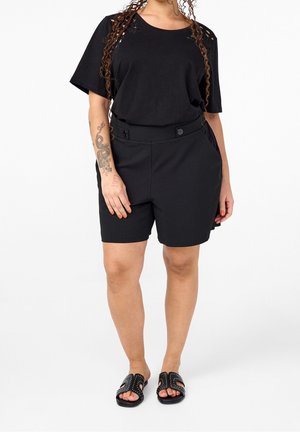 Femme aux longs cheveux bouclés portant un haut noir à manches courtes, un short noir boutonné, des sandales noires à clous, avec un grand tatouage sur l'avant-bras gauche.