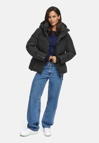 Zwarte pufferjack met een capuchon, doorstikte textuur, voorzak met ritssluiting, zijzakken, gedragen over een marineblauwe trui en gecombineerd met blauwe jeans en witte sneakers.