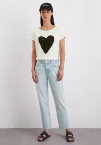Lichtgele t-shirt met een grote zwarte hartgrafiek, gecombineerd met lichtblauwe rechte spijkerbroeken en zwarte sandalen. Zwarte pet op het hoofd.