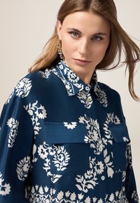 Camisa azul marino con un patrón floral blanco, diseño de botones, dos bolsillos frontales y cuello puntiagudo. Textura suave y brillante.