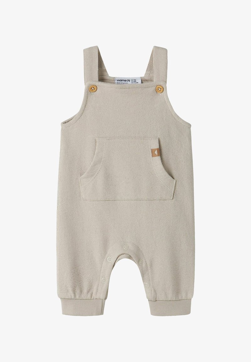 Combinaison sans manches beige pour bébé avec bretelles à boutons en bois, poche avant et pressions le long de la couture intérieure pour un change facile.