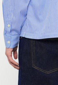 Το αριστερό χέρι ενός άνδρα χαλαρό δίπλα του, φοράει μπλε και λευκή ριγέ πουκάμισο με κουμπωμένο μανίκι και σκούρο μπλε τζιν τζιν.