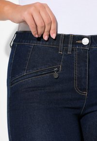 Vaqueros de mezclilla azul oscuro con un bolsillo con cremallera, costuras en contraste y un botón metálico como cierre en la cintura. Primer plano de la zona de la cintura.