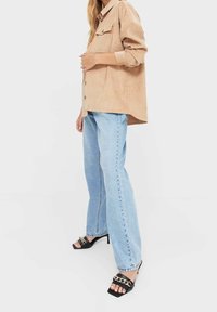 Camicia button-up in corduroy beige con taschino frontale, abbinata a jeans dritti blu chiaro e mules nere open-toe con dettaglio in catena dorata.