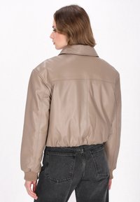 Afskåret taupe læderjakke med krave, ribbede manchetter og struktureret finish. Båret med sorte denimjeans, der viser en afslappet pasform.