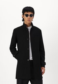 Abrigo de mezcla de lana negro con cuello alto, botones frontales y un bolsillo en el pecho. El modelo lleva gafas de sol redondas y una camiseta blanca.