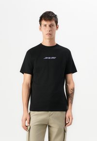 Jonge man in een zwart t-shirt met het "Santa Cruz" logo en beige cargo broek, staand met een zichtbare bloemtattoo op zijn linker onderarm.