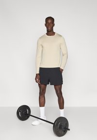 Μπεζ μακρυμάνικο πουκάμισο και μαύρο σορτς φορεμένα από ένα άτομο που ποζάρει· μπροστά βρίσκεται ένας βαράκης με δίσκους βάρους. Φοράει λευκά αθλητικά παπούτσια και κάλτσες.