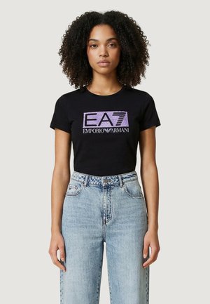 Femme aux cheveux bouclés portant un t-shirt noir EA7 Emporio Armani et un jean taille haute bleu clair, debout devant un fond uni.