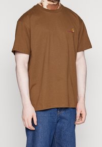 Camiseta de algodón marrón con mangas cortas, cuello redondo y logo bordado en el pecho. Combinada con pantalones vaqueros de mezclilla azul.