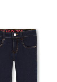 Jean en denim bleu foncé avec des coutures jaunes, featuring des poches avant et un bouton métallique de fermeture. L'intérieur de la taille présente un détail de logo rouge.