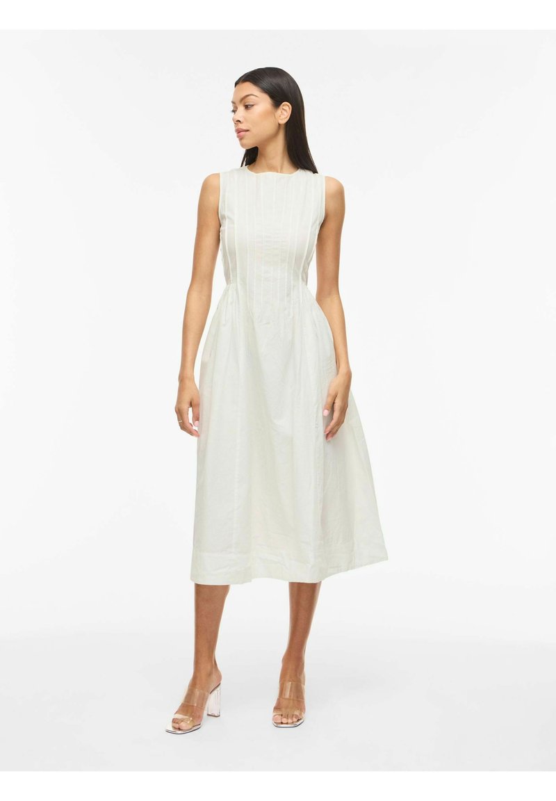 Kvinde iført hvid ærmeløs midikjole med lodrette folder, står og kigger til siden, kombineret med klare højhælede sandaler.